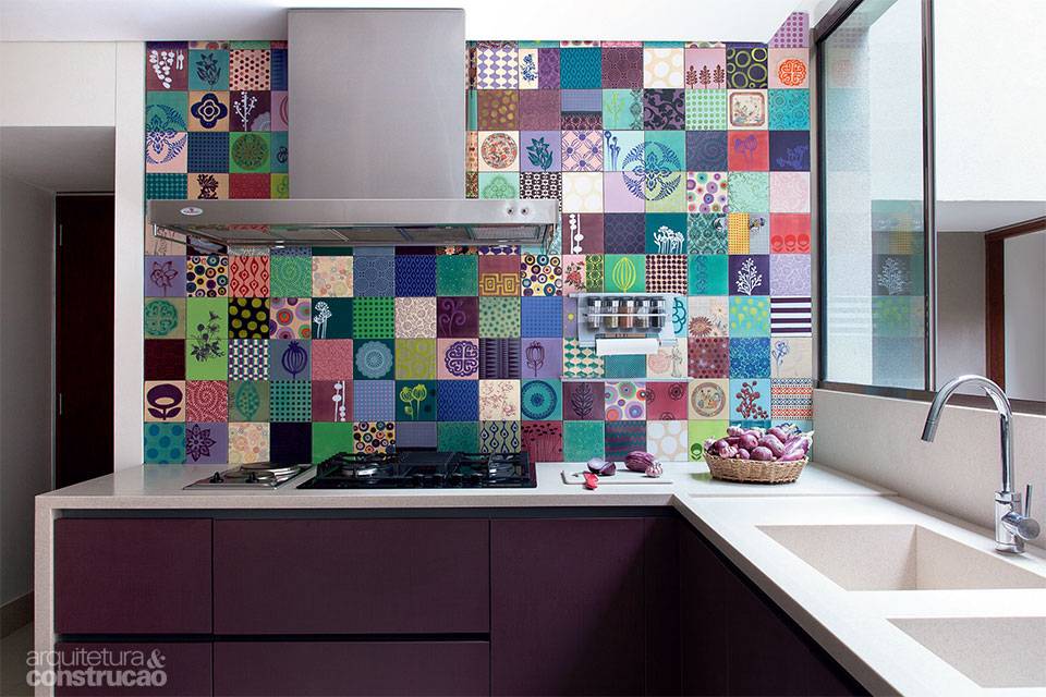 Segundo a arquiteta Katia Faria, s&atilde;o raros os clientes que d&atilde;o carta branca ao uso de cores. Livre para fazer isso neste ambiente, a profissional encontrou nos azulejos da artista Flavia Del Pra a chave para o painel de 1,40 x 2,46 m que reveste a parede atr&aacute;s do cooktop. Produzido sob encomenda, ele pautou a marcenaria que leva acabamento de laminado melam&iacute;nico berinjela (padr&atilde;o stoneware), executada pela Kitchens.