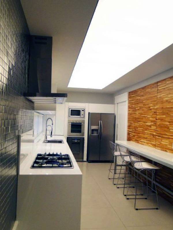 Misturar texturas tamb&eacute;m pode acontecer em cozinhas em estilo corredor. Este projeto de Michelle Donida utiliza pastilhas met&aacute;licas de um lado e madeira do outro. Para n&atilde;o sobrecarregar o ambiente, a marcenaria foi feita toda em branco.