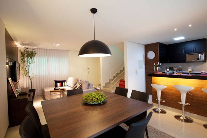 Um dos desafios da designer de interiores Sylvia Leticia de Araujo ao projetar este espa&ccedil;o, de 35,4 m&sup2;, foi integrar a cozinha com a sala de TV, o bar e a sala de jantar. Para que essa jun&ccedil;&atilde;o fosse graciosa, a profissional escolheu revestir o balc&atilde;o da cozinha com painel amadeirado, que se assemelha &agrave; decora&ccedil;&atilde;o do resto do ambiente.