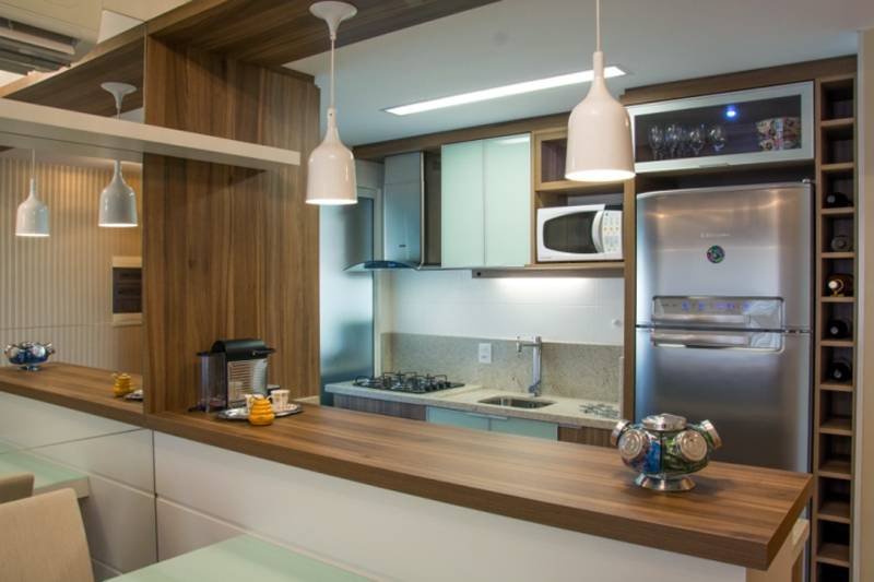 A cozinha integrada assinada por Camila Chalon utiliza marcenaria sob para os arm&aacute;rios e a bancada (do outro lado, h&aacute; um pequeno arm&aacute;rio que fica escondido). Para aproveitar melhor o espa&ccedil;o, o v&atilde;o entre a parede e a geladeira se transformou em adega para bebidas.