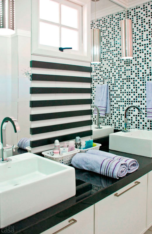 A bancada deste banheiro se vale da beleza do granito preto s&atilde;o gabriel. Sobre ele, v&atilde;o duas cubas de apoio, retas e elegantes. Ripas decorativas de MDF escurecido foram fixadas na parede entre as pias. Para completar o visual, a superf&iacute;cie na lateral da bancada recebeu que imita uma miscel&acirc;nea de pastilhas.