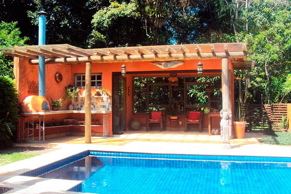 O pergolado de eucalipto tratado com policarbonato suspenso por pinos niveladores protege a &aacute;rea gourmet r&uacute;stica. Com forno a lenha e parede revestidos com terra pela t&eacute;cnica de pau a pique. O mosaico no piso &eacute; feito de ladrilho hidr&aacute;ulico executado na obra e sobrep&otilde;e a bancada de concreto, cuba de cobre e torneira com conex&otilde;es de cano de cobre. Projeto de Pedrinha Parisi.
