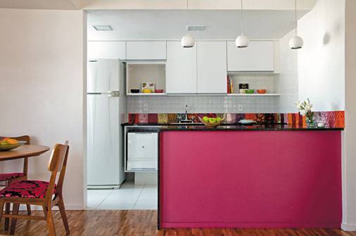 Neste apartamento, a reforma da cozinha trouxe jovialidade e amplitude para a &aacute;rea social. Totalmente branca, a decora&ccedil;&atilde;o permitiu a ousadia do tom rosa da bancada. Para quebrar o impacto, um tampo de granito preto &eacute; colocado, ao lado de azulejos coloridos.