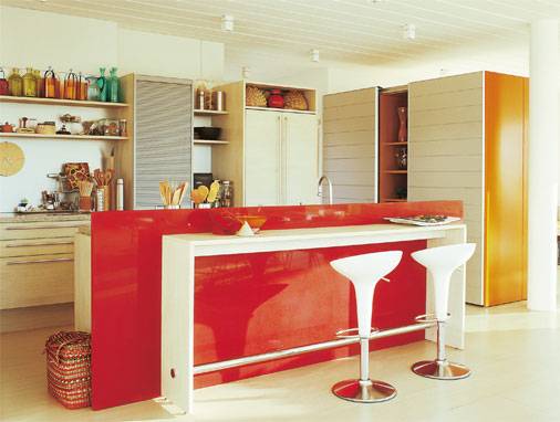 A integra&ccedil;&atilde;o desta cozinha &eacute; diferente: foi feita com o quarto de um casal. A proposta pede um material aconchegante, por isso a madeira foi utilizada estruturalmente. A bancada, que separa visualmente os espa&ccedil;os, &eacute; camuflada por uma placa vermelha feita de p&oacute; de quartzo e granito processado.