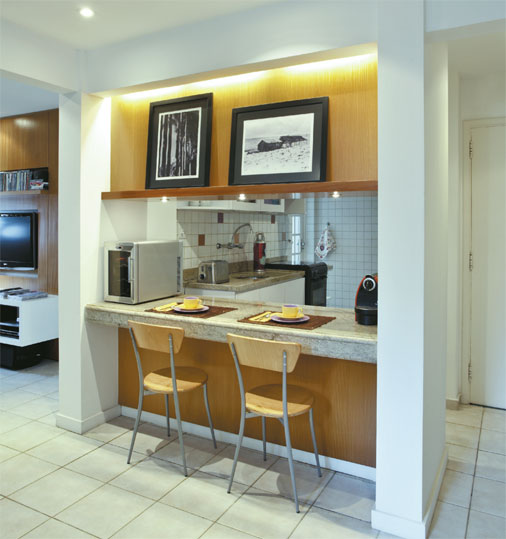 Para deixar a cozinha integrada mais elegante, a arquiteta Carola Gouv&ecirc;a fechou as laterais da bancada. As novas colunas de MDF s&atilde;o facilmente desmont&aacute;veis e o morador pode lev&aacute;-las caso queira sair deste apartamento alugado. "Era importante que tudo fosse facilmente desmont&aacute;vel e reaproveit&aacute;vel para o caso de mudan&ccedil;a", lembra a arquiteta. Um painel de peroba superior liga as colunas e possui um nicho para apoiar quadros. Na parte inferior, a mesma madeira cria a unidade de toda a reforma.