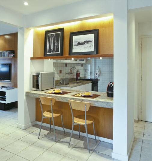 Para deixar a cozinha integrada mais elegante, a arquiteta Carola Gouv&ecirc;a fechou as laterais da bancada. As novas colunas de MDF s&atilde;o facilmente desmont&aacute;veis e o morador pode lev&aacute;-las caso queira sair deste apartamento alugado. "Era importante que tudo fosse facilmente desmont&aacute;vel e reaproveit&aacute;vel para o caso de mudan&ccedil;a", lembra a arquiteta. Um painel de peroba superior liga as colunas e possui um nicho para apoiar quadros. Na parte inferior, a mesma madeira cria a unidade de toda a reforma.