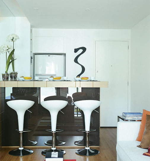 A solu&ccedil;&atilde;o para o pouco espa&ccedil;o, neste apartamento de 50 m&sup2;, foi criar uma cozinha integrada. A pia ganhou lugar atr&aacute;s do balc&atilde;o revestido de vidro espelhado. Prateleiras e arm&aacute;rios foram feitos ao lado do nicho da geladeira.