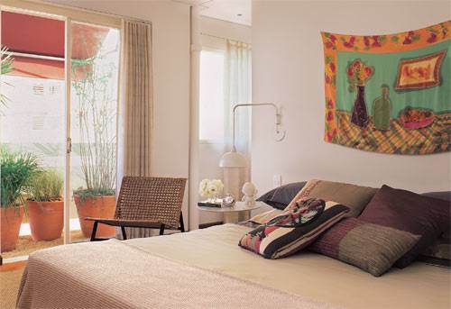 Tons terrosos e arroxeados predominam nas almofadas (Benedixt, Emp&oacute;rio Beraldin e Esencial) sobre a cama, vestida com enxoval da Blue Gardenia. Na parede, seda pintada pela amiga Andrea Kramer. De linhas elegantes, a cadeira de fibra e metal foi comprada na loja A Lot Of.