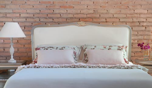 O contraste entre a parede r&uacute;stica de tijolos &agrave; vista e as linhas cl&aacute;ssicas dos m&oacute;veis cria uma agrad&aacute;vel surpresa neste quarto de casal. O ponto alto do ambiente &eacute; a cabeceira (modelo Nice, 1,60 x 1,35 m de altura. Pre&ccedil;o: 8,4 mil reais) da Madeira Bonita. A pe&ccedil;a de madeira entalhada e acabamento dourado &eacute; estofada e revestida de sarja e vem com um suporte pr&oacute;prio de fixa&ccedil;&atilde;o. Nas laterais, foram instaladas as duas mesinhas redondas e a c&ocirc;moda, com tampo e frente de gavetas revestidas de espelho bisotado.
