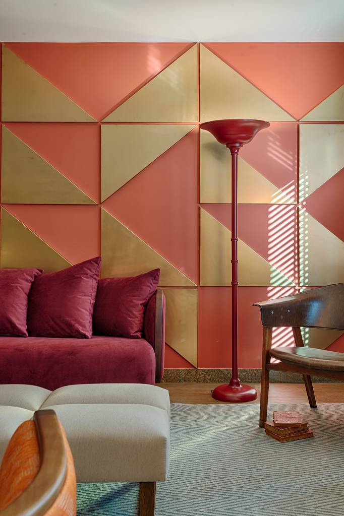 Esta sala, criada pela Melina Mundim, trabalha principalmente com tons terrosos e avermelhados, com exce&ccedil;&atilde;o das cadeiras ao fundo, dos bancos, do tapete e dos pain&eacute;is triangulares. A decora&ccedil;&atilde;o tamb&eacute;m possui uma influ&ecirc;ncia retr&ocirc;, como pode ser observado na lumin&aacute;ria de ch&atilde;o vermelha, nos formatos arredondados do sof&aacute; e das poltronas e na estampa geom&eacute;trica criada pelos pain&eacute;is dourados na parede.