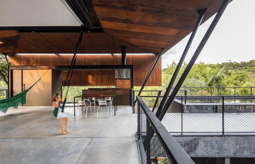 A casa é formada por dois volumes estreitos empilhados um sobre o outro para formar uma forma em T. Paredes de vidro deslizantes foram instaladas ao redor da estrutura do piso superior para permitir que os espaços principais se abram ao ar livre e ofereçam ventilação cruzada. Um telhado de aço de intemperismo cobre a estrutura e é dividido em dois, permitindo que uma brisa passe pela casa. A casa é formada por dois volumes estreitos empilhados um sobre o outro para formar uma forma em T. Paredes de vidro deslizantes foram instaladas ao redor da estrutura do piso superior para permitir que os espaços principais se abram ao ar livre e ofereçam ventilação cruzada. Um telhado de aço de intemperismo cobre a estrutura e é dividido em dois, permitindo que uma brisa passe pela casa.