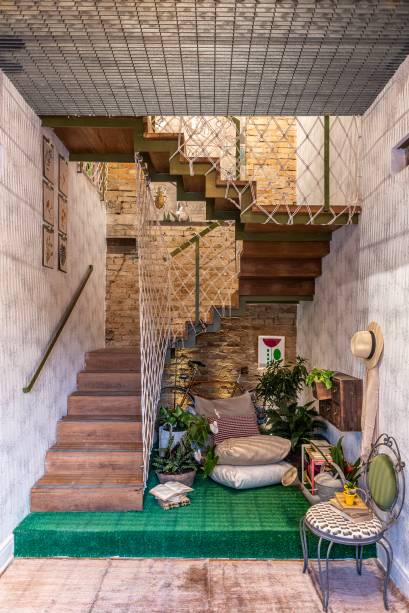 Hall de entrada e Bilheteria - Isabela Scaff e Maria Gabriela Azevedo, e paisagismo de Clariça Lima. Com 15 m² e pé-direito duplo, o hall é aberto para os dois andares da casa. O rústico ganha espaço na ambientação, como nas cordas que entrelaçam o guarda-corpo original em ferro. No vão, o mobiliário convive com plantas e flores, além do tapete que imita grama.