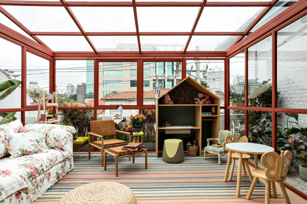 Varanda - Marina Linhares. Um gazebo de 39m&sup2;, constru&iacute;do a partir de uma estrutura met&aacute;lica e em vidro, explorando a luz natural e integrado ao paisagismo de Gabi Pileggi, do Jardineiro Fiel. Pensado para reunir a fam&iacute;lia em um momento de contempla&ccedil;&atilde;o do horizonte, o espa&ccedil;o &eacute; pontuado por flores e plantas. O interior apresenta m&oacute;veis confort&aacute;veis estofados com tecidos naturais e objetos de design. Uma estante com dois metros e formato de casinha abriga uma cole&ccedil;&atilde;o de brinquedos.