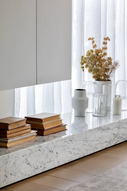 O armário branco, feito para criar o efeito de “estar voando”, ganha o contraponto com o grande bloco de mármore carrara que em sua composição traz as cores do ambiente como num espelho das nuvens.