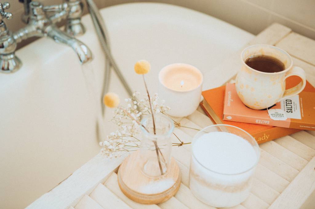 spa-em-casa-maddi-bazzocco- Velas, flores, livros e uma caneca de chá sobre um apoio de madeira em uma banheira de banho