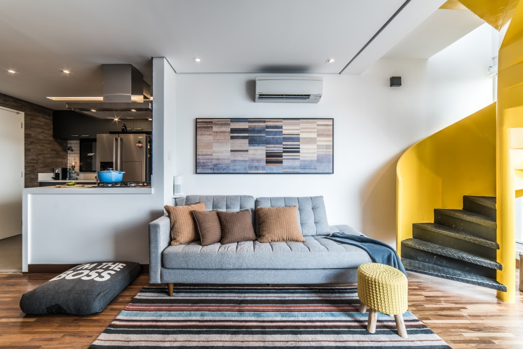 8-dicas-essenciais-para-adaptar-a-decoracao-da-casa-com-bicho-de-estimacao-casa.com (3) Sala de estar com um sofá cinza e almofadas marrons. Acima dele, um quadro com retângulos nas cores azul, marrom, bege e preta. No chão, um tapete listrado e um banco com pés de madeira e capa de crochê amarelo. Ao lado esquerdo da imagem, uma cama de cachorro onde se lê "I'm the boss" (Eu sou o chefe, em inglês). No mesmo lado, ao fundo, uma bancada com cooktop e panela azul, e mais atrás a cozinha. Do lado direito, uma escada de metal, com os degraus em preto e guarda-corpo em amarelo.
