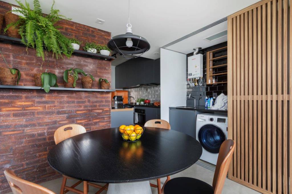 SP Studio_Maxhaus_(RafaelRenzo)-36 Área de receber com mesa e cadeiras em madeira. Parede de tijolos com prateleiras com vasos de plantas. Porta de madeira deslizável que abriga máquina de lavar e pia.