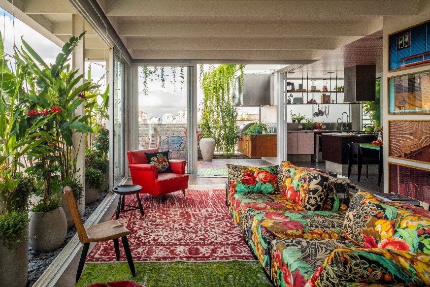 apartamento-julliana-camargo-colorido-14 Estar com tapete vermelho sobre tapete verde. Sofá florido e poltrona vermelha. Folhagens altas na parede da esquerda. Trepadeiras ao fundo