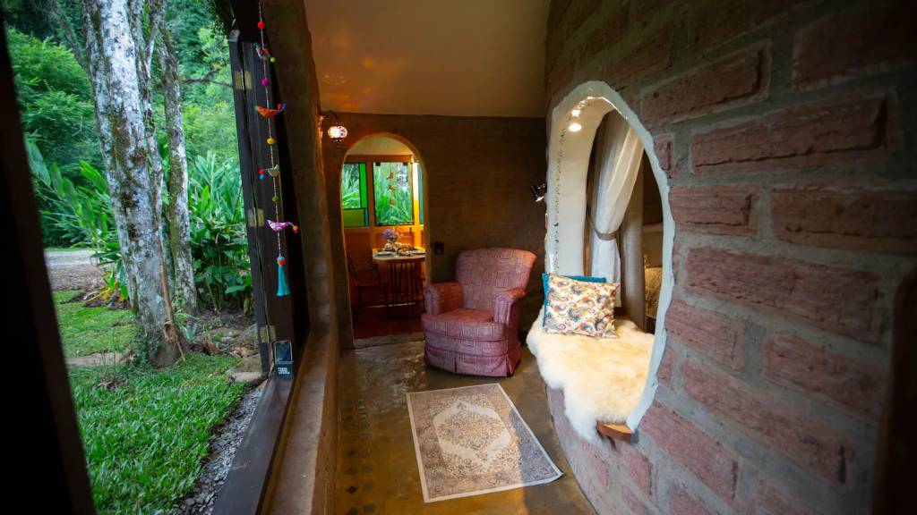 casas-tematicas-do-airbnb-no-brasil-hobbits-herois-e-amantes-de-vinho-casa.com-1-hobbit Hall de entrada de tijolos laranjas, com uma poltrona vermelha, um tapete estampado e um peitorial da janela arredondado com almofadas para sentar.
