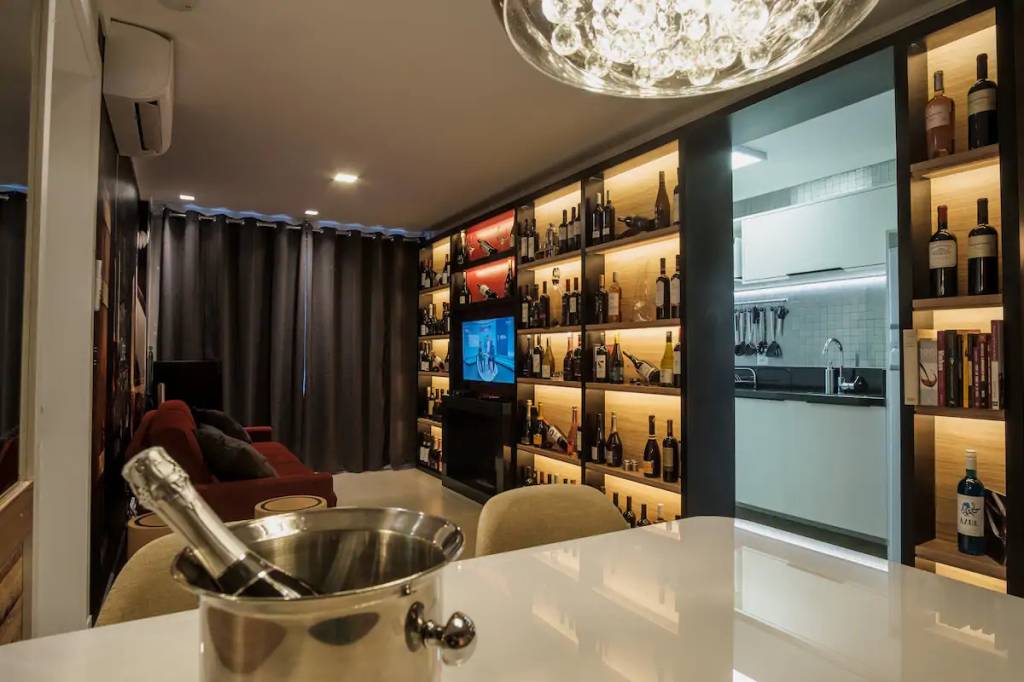 casas-tematicas-do-airbnb-no-brasil-hobbits-herois-e-amantes-de-vinho-casa.com-3-vinhos Sala de estar, com estante cheia de vinhos nos nichos. No centro, uma TV é emoldurada por eles.