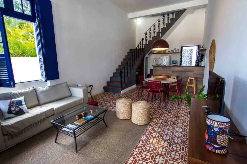 casas-tematicas-do-airbnb-no-brasil-hobbits-herois-e-amantes-de-vinho-casa.com-4-alceu-valenca Sala com cozinha integrada. À esquerda, um sofá cinza com uma alomfada estampanda com o rosto do cantor Alceu Valença. À frente dela, uma mesa de centro com tampo de vidro, doi pufes beges, um tapete estampando Ao fundo, do lado esquerdo uma escada preta, ao lado, no andar inferior, uma mesa com cadeira vermelhas, a bancada da cozinha em último plano, os eletrodomesticos