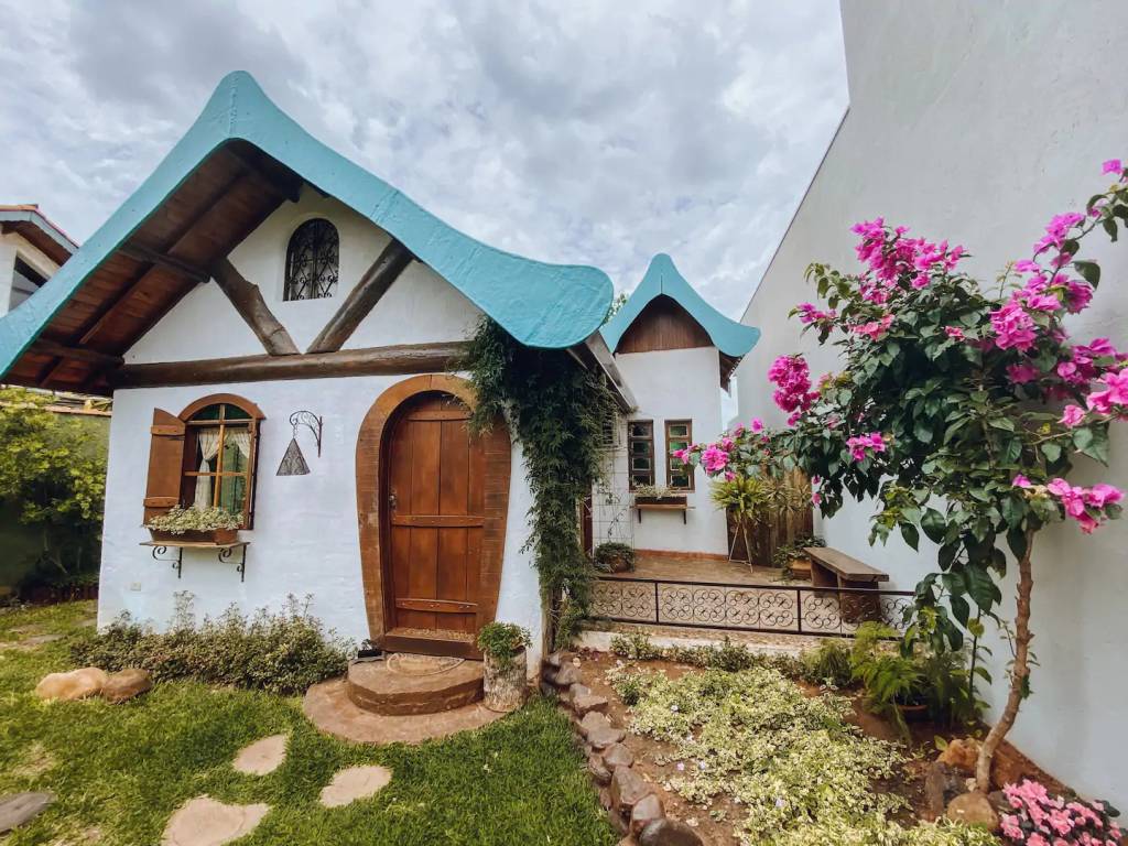 casas-tematicas-do-airbnb-no-brasil-hobbits-herois-e-amantes-de-vinho-casa.com-5-conto-de-fadas Casa de conto de fadas com parede branco, teto triangular de madeira, porta e janela com batente arredondado