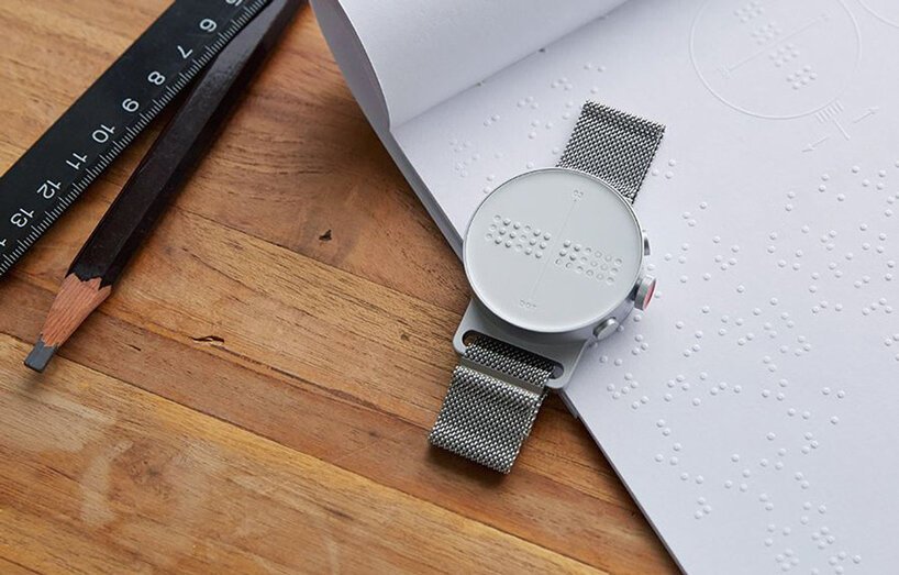 dot-watch-relogio-braille-designboom-01 Dot watch sobre mesa de madeira e cadeno braille. No canto superior esquerdo, um lápis preto e uma régua