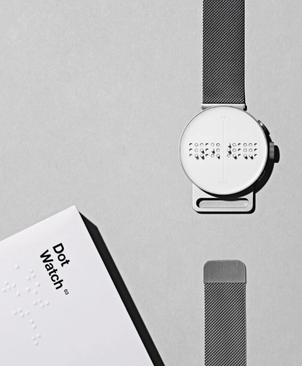 dot-watch-relogio-braille-designboom-02 Dot Watch com a pulseira aberta em uma das extremidades em fundo cinza. No canto inferior esquerdo, manual do relógio em braille