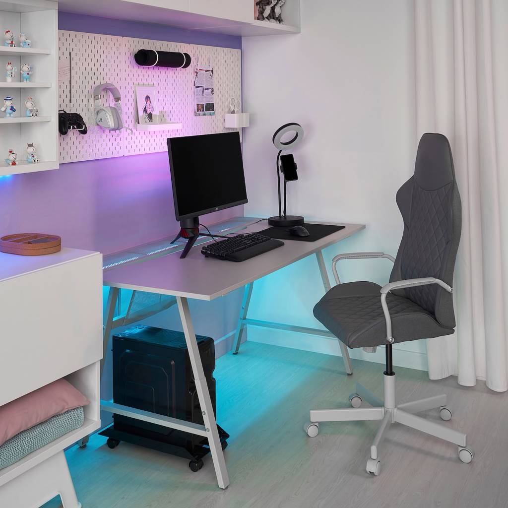 ikea-lanca-colecao-de-moveis-para-e-sports-casa.com-3 Mesa cinza, com luzes lilás na parte traseira superior e azul claro na parte inferior. Um monitor, telcado e mous sobre a mesa. Na parede, um mural branco com fone de ouvido, controle de video game e um porta retrato. A CPU fica no chão, do lado esquerdo da mesa, e a cadeira é cinza.