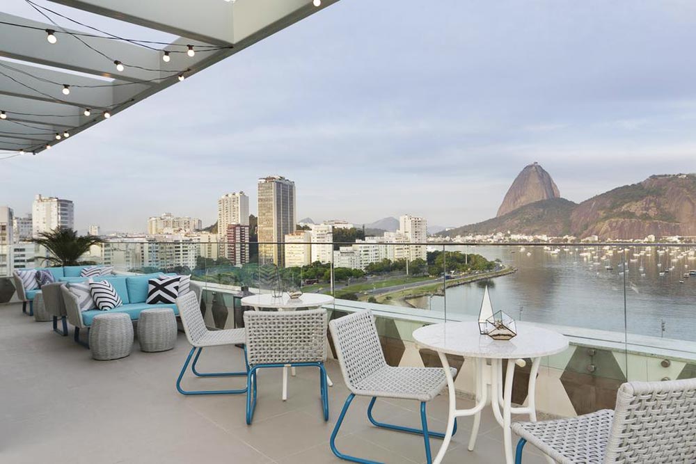 rooftop-a-tendencia-na-arquitetura-contemporanea-yoo2-casa.com-1 Cadeiras e mesas brancas no Rooftop do Yoo2, ao Fundo, a cidade do Rio De Janeiro, com o Pão de Açúcar e o mar.