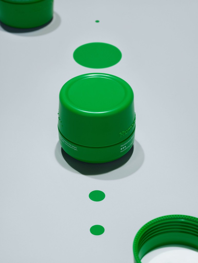 Produto de cuidado facial, com embalagem verde, cilíndrica em fundo branco azulado