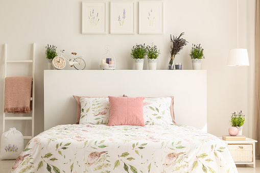 Pink pillow on bed with headboard in white bedroom interior with plants and posters. Real photo