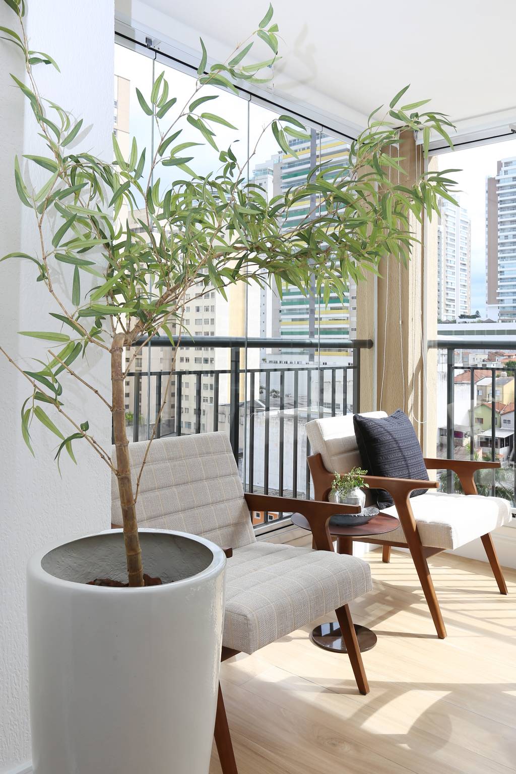 No apartamento de 65m&sup2;, a varanda integrada combina o tom amadeirado das poltronas, o porcelanato que simula os veios da madeira, e a luz solar que adentra o ambiente por conta do fechamento de vidro. O clima de paz e contato com a natureza &eacute; completada com o vaso cer&acirc;mico com planta. Um pequeno o&aacute;sis em meio ao arranha-c&eacute;u de S&atilde;o Paulo | Projeto Oliva Arquitetura