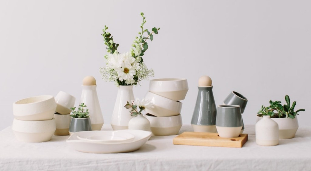 Mesa com pratos, copos e tigelas em cerâmica branca. Pequenas folhas e flores nos vasos. 