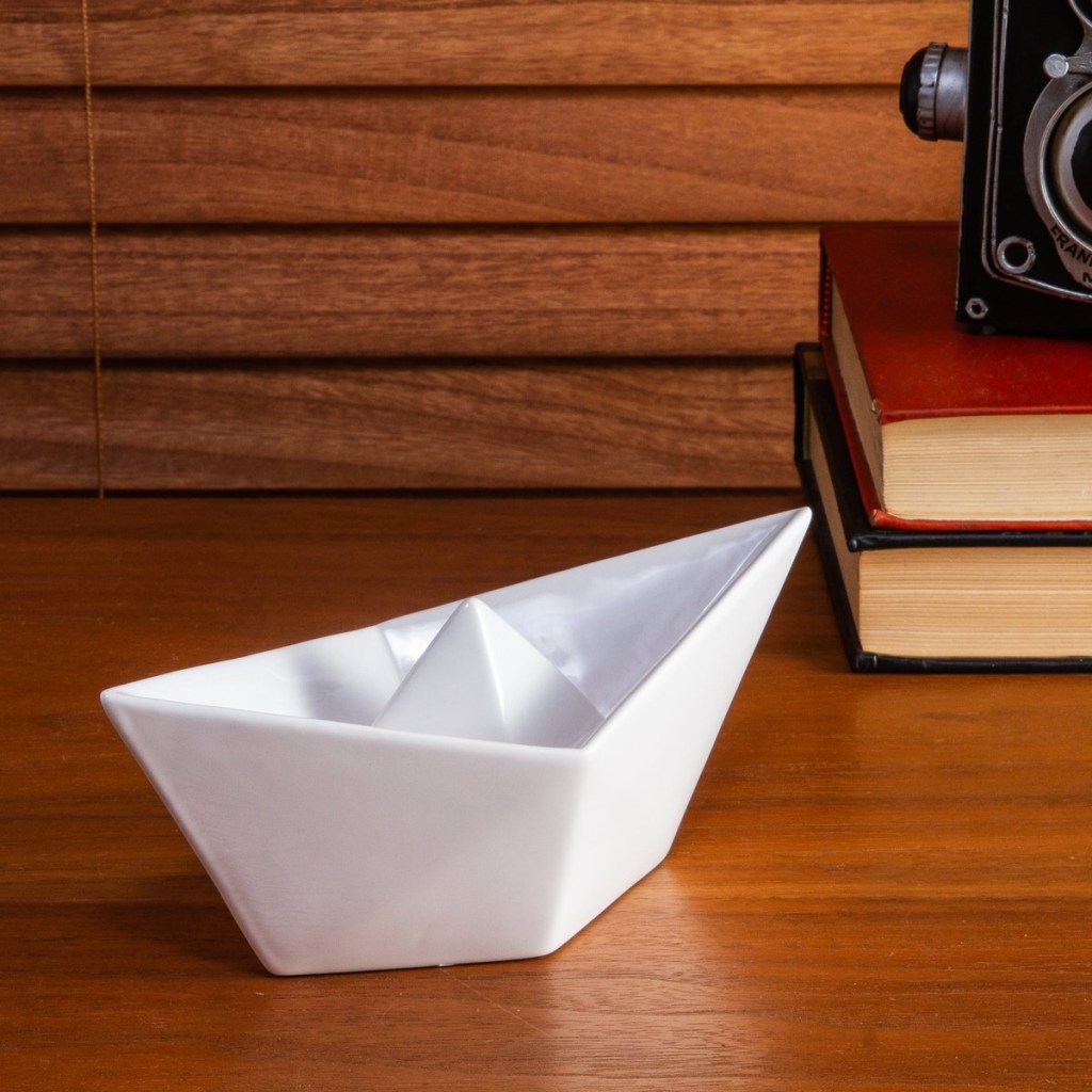 Barco de papel de cerâmica, sobre bancada de madeira com livros e máquina fotográfica antiga ao fundo