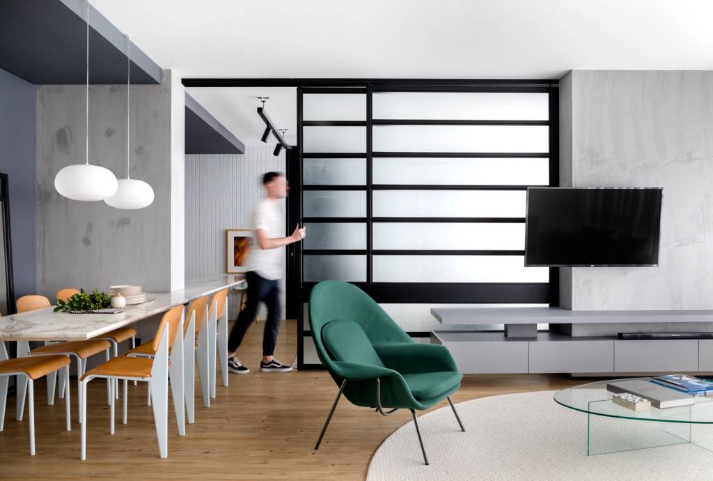 estudio-de-73-m²-com-planta-integrada-e-design-moderno-casa.com-archdaily-studio-gabirel-bordin-fabio-junior-severo-1 Sala de estar com piso de madeira,com tapete redondo branco no centro,sob sofá da mesma cor. Ao lado, uma poltrona verde, com uma mesa de janta branca, com detalhes em madeira. Ao fundo, um jovem rapaz emourra uma porta de correr que separa a área social da área íntima
