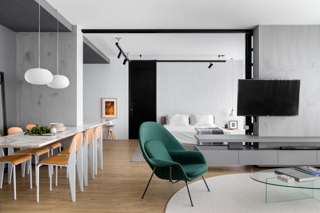 estudio-de-73-m²-com-planta-integrada-e-design-moderno-casa.com-archdaily-studio-gabirel-bordin-fabio-junior-severo-6 Sala de estar com piso de madeira,com tapete redondo branco no centro,sob sofá da mesma cor. Ao lado, uma poltrona verde, com uma mesa de janta branca, com detalhes em madeira. Ao fundo, a porta de correr aberta mostra as áreas social e íntima integradas