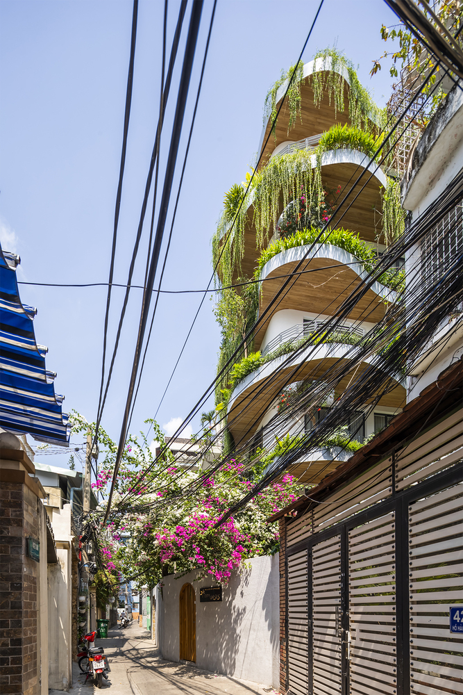 Visão da fachada, com prédios menores ao lado, com vista para as varandas com plantas tropicais no prédio branco e com detalhes em madeira clara