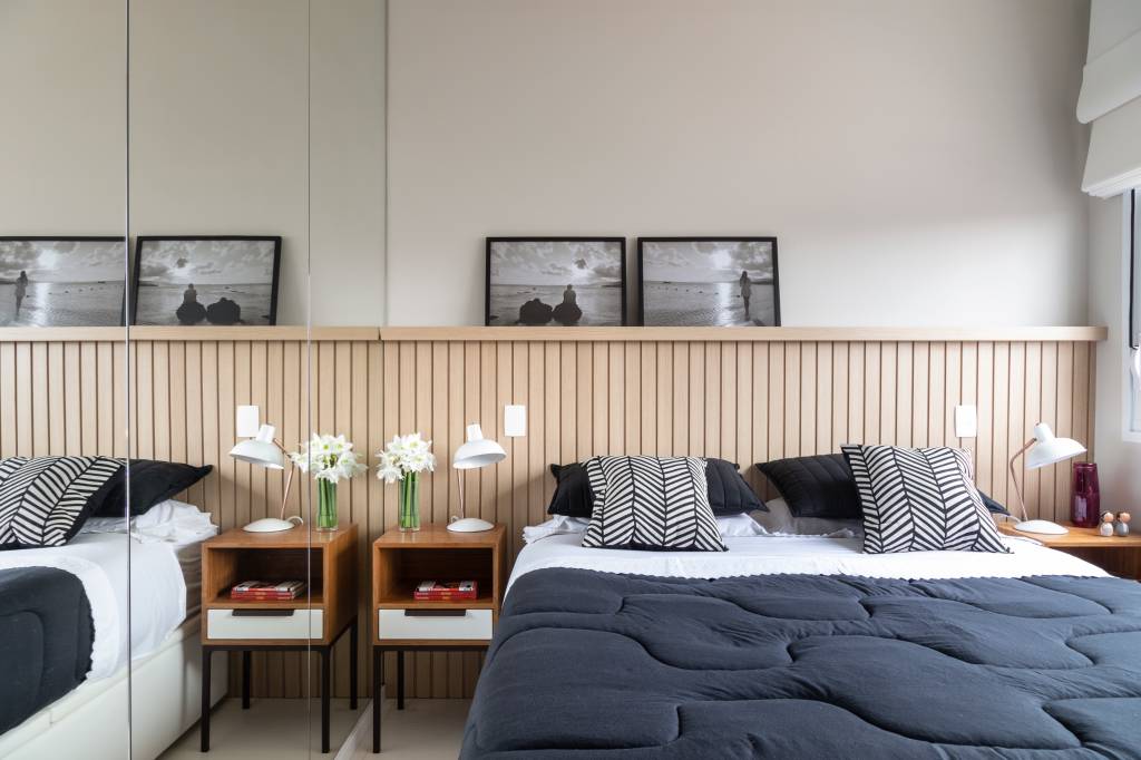 modernidade-e-leveza-no-decor-casa.com _MG_2079 Quarto do casal, com cabeceira de madeira ripada, doi quadros em preto e branco acima dela. Na lateral da cama, uma mesa de madeira com uma gaveta branca, livros no nicho pequeno e flores sobre ela. Na cama, o edredom é azul escuro. Na lateral, um espelho de fora a fora esconde o guarda roupas