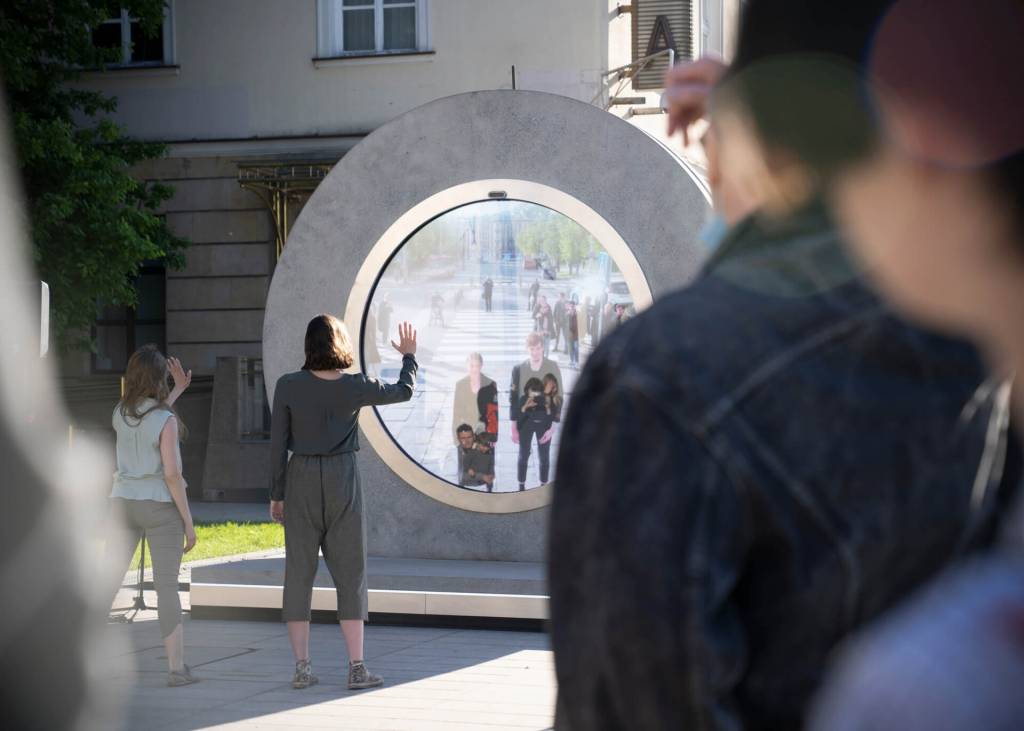 portal-permite-ver-outra-parte-do-mundo-em-tempo-real-casa.com-3 Círculo com tela no centro que permite ver outro lugar do planeta. Atrás, uma grande construção. À frente, diversas pessoas olham através do portal e acenam para o outro lado
