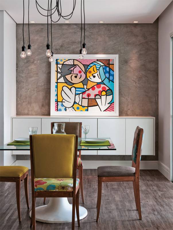 A parede da sala de jantar deste apartamento recebeu cimento queimado comum com uma dem&atilde;o de resina sint&eacute;tica. O ar r&uacute;stico &eacute; quebrado pela tela colorida. O tampo de vidro incolor leva adesivo vin&iacute;lico na face de baixo. Projeto de Cec&iacute;lia Guerra e Cristiane Tamburri.