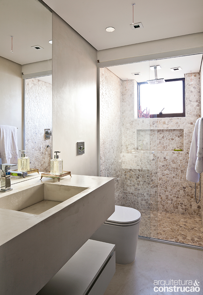 Com a ideia de simular um banho em meio &agrave; natureza, seixos da linha Mosaico Telado &Ocirc;nix (Palimanan) recobrem paredes e piso do boxe no projeto do escrit&oacute;rio H2C Arquitetura. No mesmo tom, o ciment&iacute;cio Tecnocimento Classic cinza-claro (NS Brazil) forra o resto do banheiro.