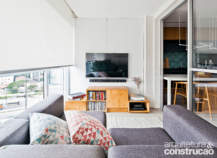 Com piso nivelado e igualado ao restante do apartamento e fechamento de vidro, o terra&ccedil;o virou home theater. O painel de madeira com laminado branco recebeu a TV e embutiu tomadas e fia&ccedil;&atilde;o. A tela solar da Hunter Douglas (Arthur Decor) regula a luminosidade. Almofadas da Cremme e tapete da Phenicia Concept.