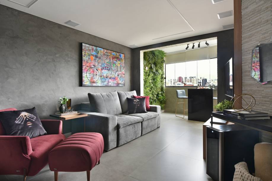 Deste ângulo, é possível visualizar a área da sala de estar/home theater e a varanda integrada. Com a predominância do cinza, os móveis e outros elementos do décor, são os responsáveis por evidenciarem os pontos de cor