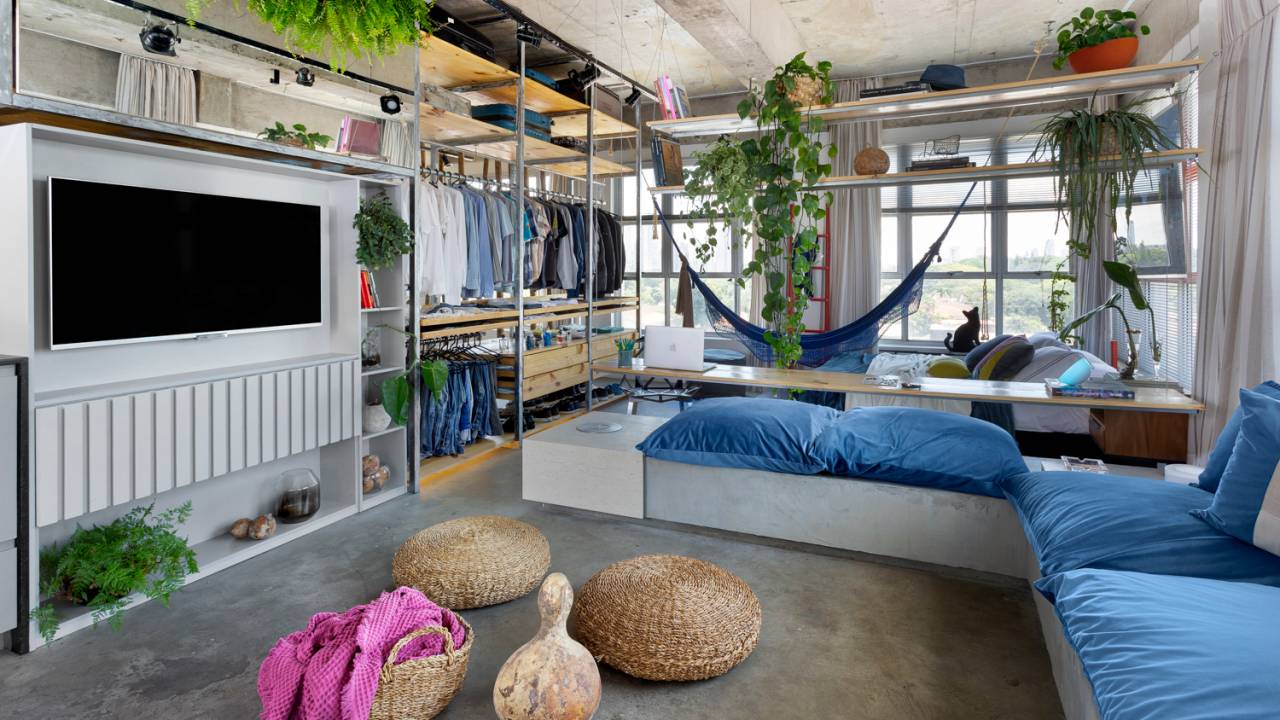 Sala de estar com sofá de alvenaria, integrada com quarto, prateleiras permeáveis com closet e vasos de planta. Cestarias em primeiro plano.