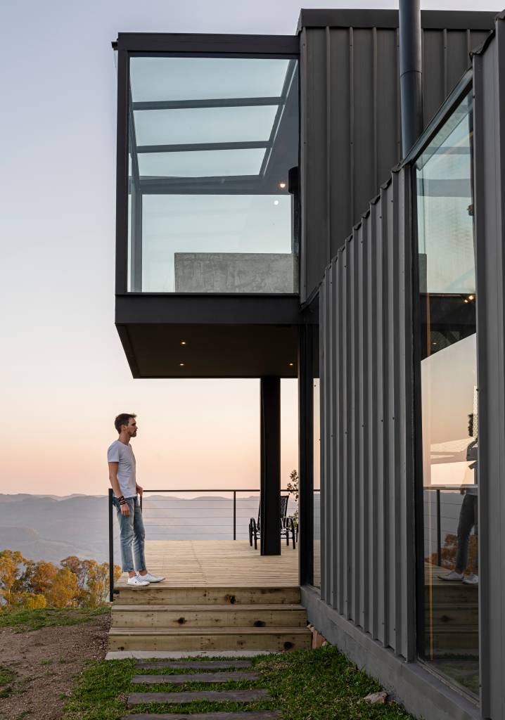 cabana-contemporanea-glamping-caxias-do-sul-guilherme-jordani-fachada-cabana-steel-frame-natureza-mirante Homem aprecia vista de montanha a partir do deck de madeira de sua cabana em steel frame.