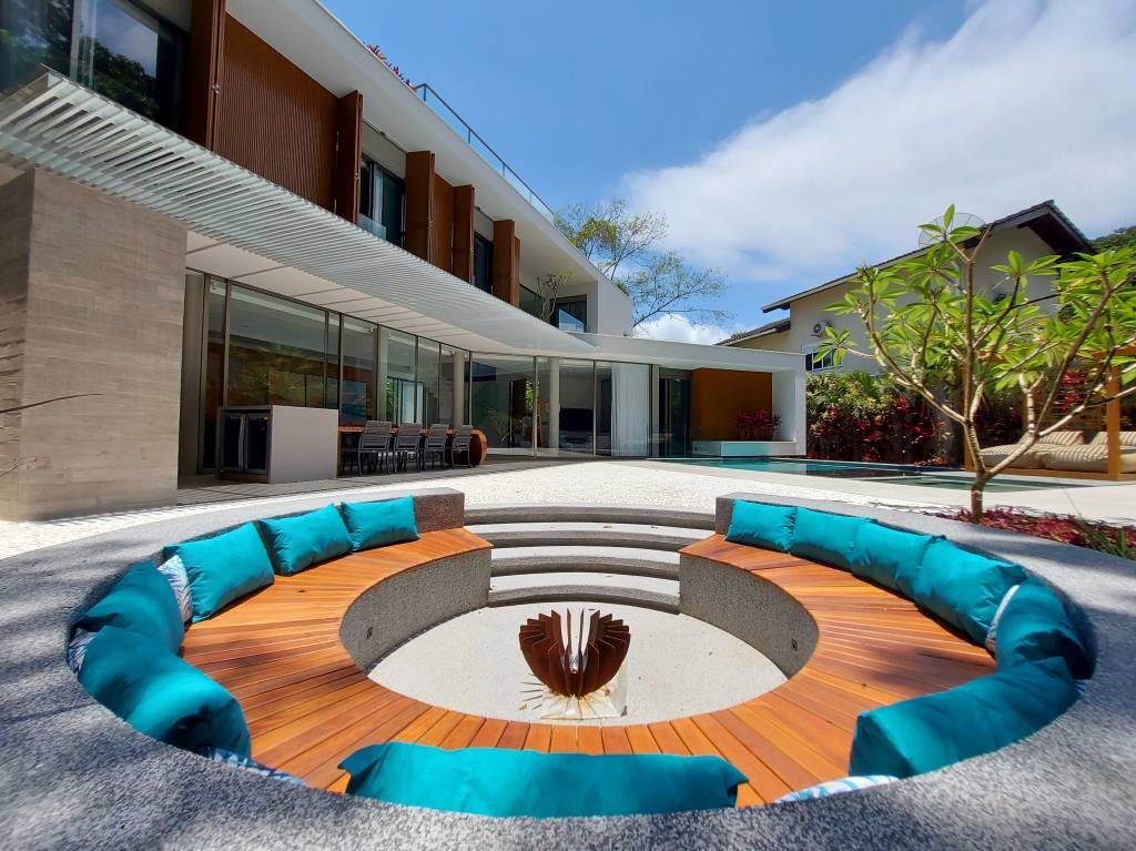 casa-na-praia-para-2-casais-de-amigos-cristiano-cabral-praca-do-fogo-area-externa-varanda-jardim Em primeiro plano vemos um círculo semienterrado com banco e almofadas para sentar. Ao centro, um ponto para uma fogueira ao ar livre. Ao fundo, a fachada interna da casa, com varanda e janelas grandes.