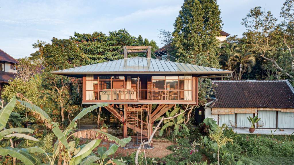 conheca-casa-na-arvore-em-bali-arley-mardo-fachada-com-escada-de-acesso-jardim Imagem mostra casa na árvore suspensa do solo com escada de acesso e jardim ao redor.