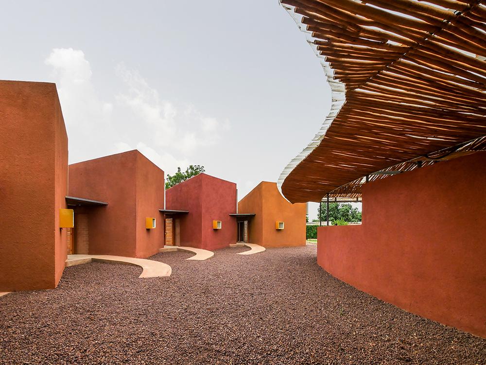 francis-kere-vencedor-pritzker-2022-divulgacao-leo-doctors-house Imagem mostra fachada do projeto Leo Doctors Housing, construção de terra pintada em cores naturais, chão de pedras e cobertura de bambu.