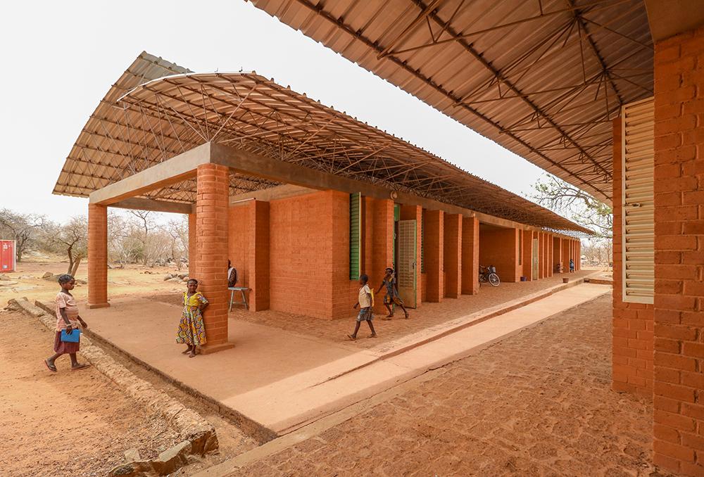 francis-kere-vencedor-pritzker-2022-divulgacao-opera-village Imagem mostra prédio horizontal com tijolos aparentes, cobertura com telhas metálicas e algumas crianças em frente, em paisagem árida africana